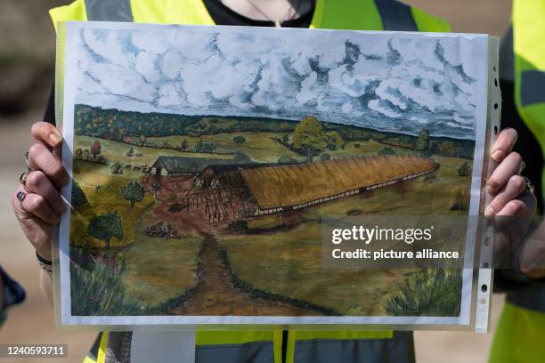 May 2022, Lower Saxony, Göttingen: An excavation worker shows a sketch with a Neolithic longhouse at the archaeological site. The electricity grid...