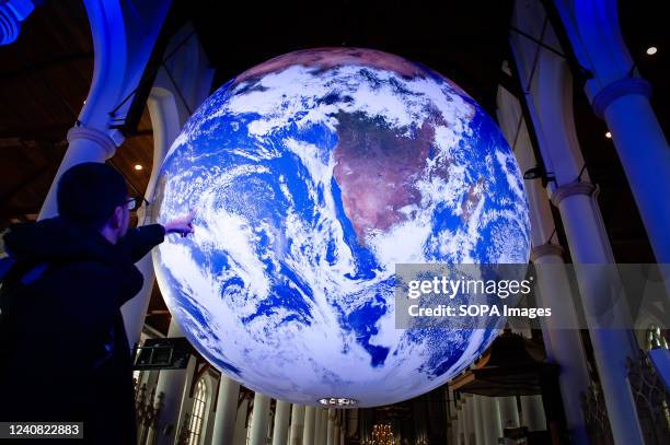 Man is seen pointing at a big Earth ball placed inside of a church. Gaia is a touring artwork by UK artist Luke Jerram. Measuring seven meters in...