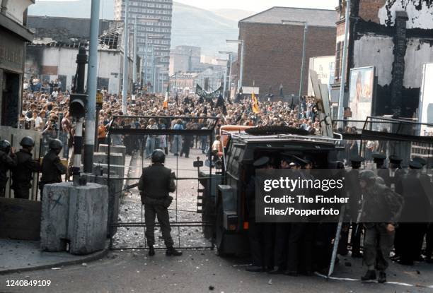 British troops and Irish police stand behind a barricade and a row of shields against rioters during The Troubles in the city of Belfast, Northern...