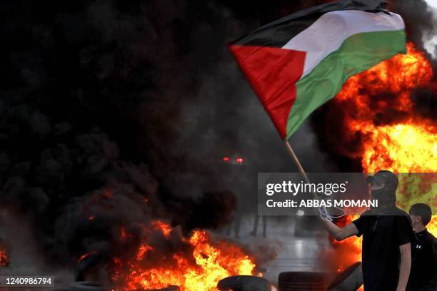 Masked protester waves a Palestinian flag near a tire fire during clashes with Israeli forces following a protest to denounce the annual nationalist...