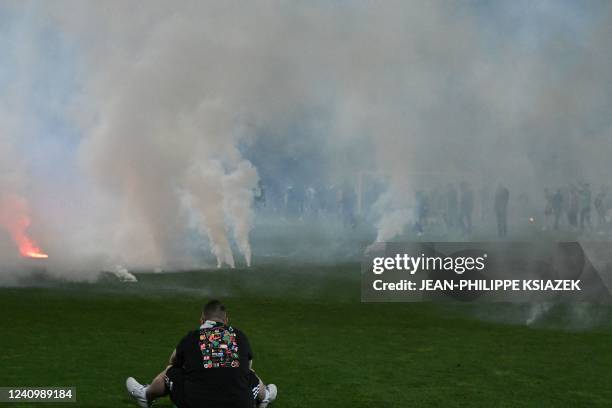 Football fan sits on the pitch as riot police officers in background clash with Saint-Etienne's fans who invaded the pitch after being defeated by...