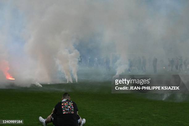Football fan sits on the pitch as riot police officers in background clash with Saint-Etienne's fans who invaded the pitch after being defeated by...