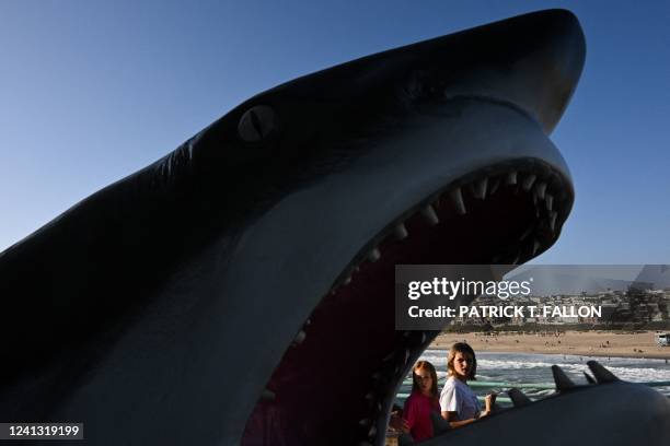 People carry ice cream cones as they walk past a statue of a great white shark on the Manhattan Beach pier on June 14, 2022 in Manhattan Beach,...