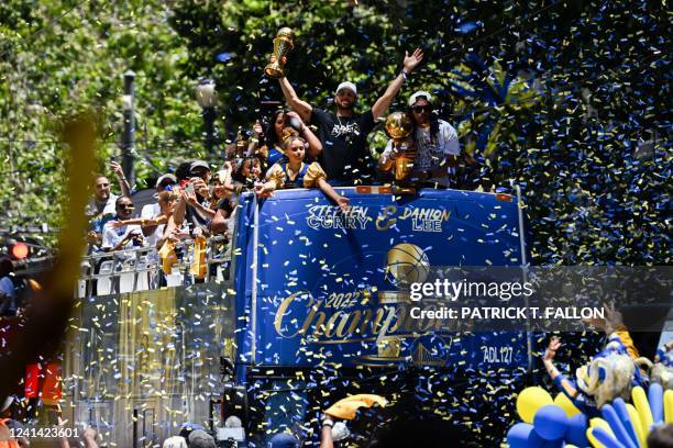 Stephen Curry and Damion Lee hold trophies as they wave from a confetti-covered double-decker bus as basketball fans cheer them on during the Golden...