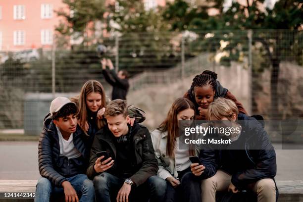 multi-ethnic teenage friends using smart phones while sitting at park in city - teenagers only stock pictures, royalty-free photos & images