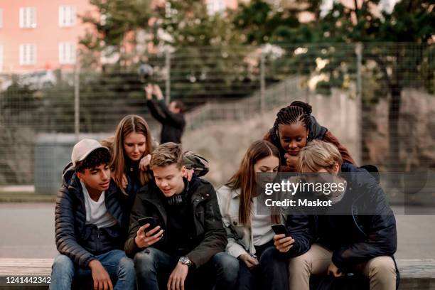 multi-ethnic teenage friends using smart phones while sitting at park in city - grupo de adolescentes imagens e fotografias de stock