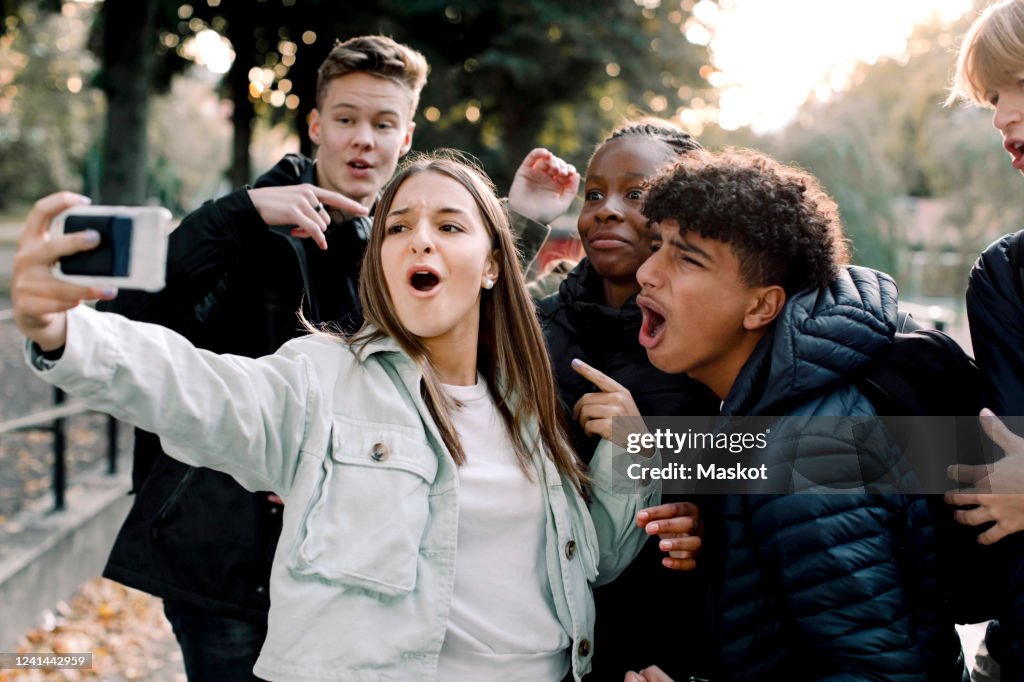 Cheerful teenage friends taking selfie through mobile phone while standing on street in city