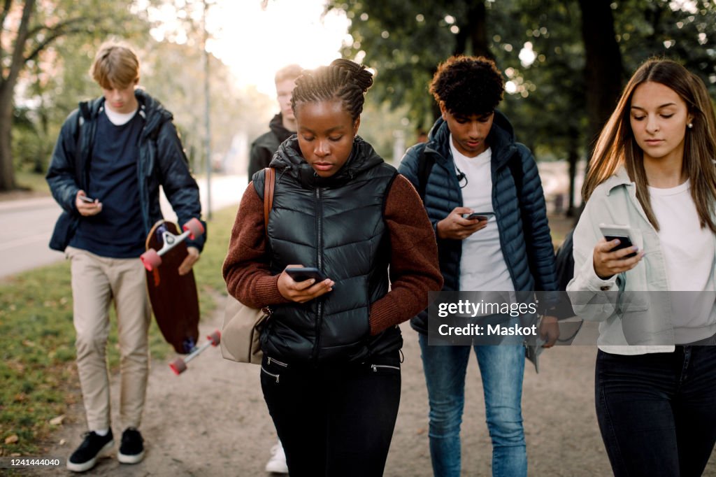 Multi-ethnic teenage friends using smart phones while walking on street in city