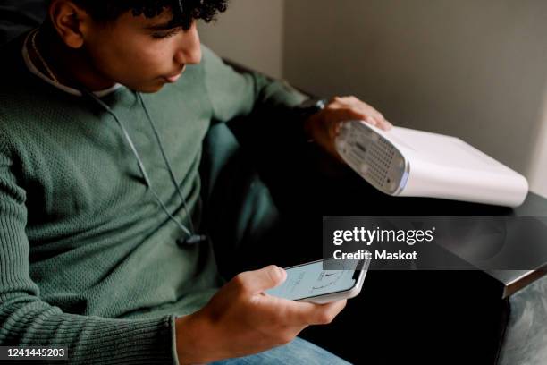 teenage boy holding router while checking wi-fi connection on mobile phone at home - wifi stockfoto's en -beelden