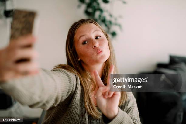 teenage girl showing peace sign while taking selfie through mobile phone at home - girl selfie fotografías e imágenes de stock
