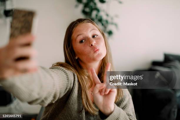 teenage girl showing peace sign while taking selfie through mobile phone at home - female puckered lips stockfoto's en -beelden