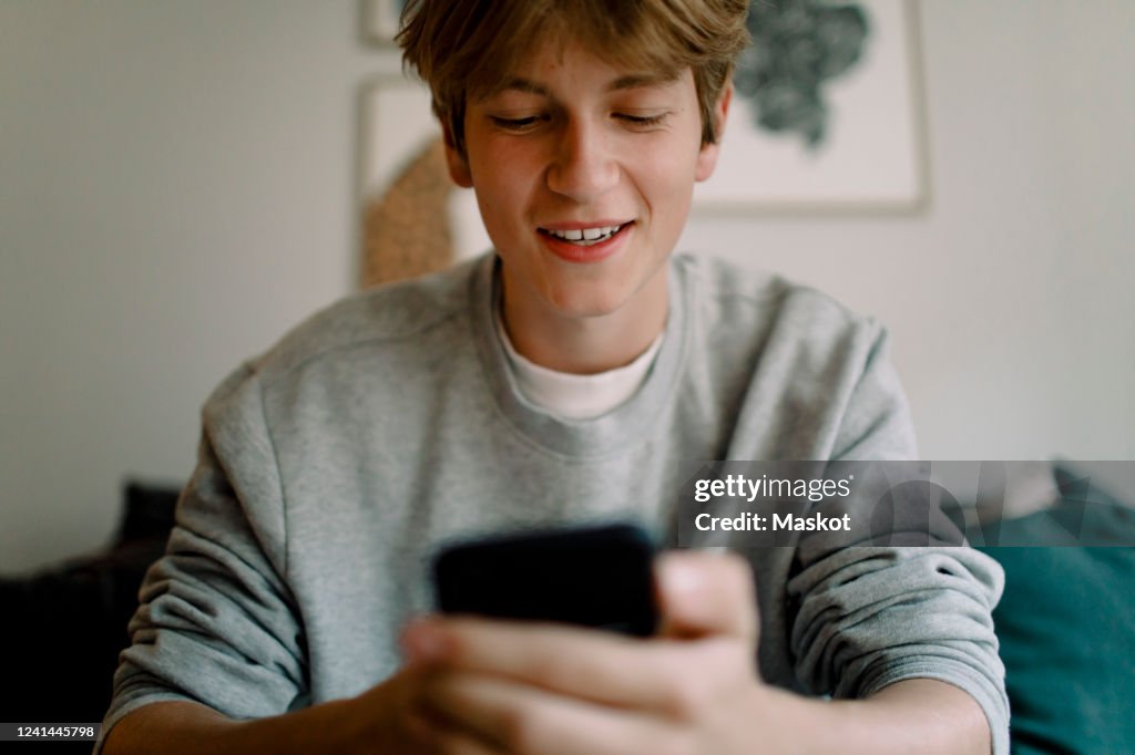 Smiling teenage boy using smart phone while sitting at home
