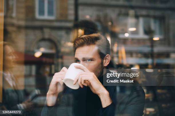 businessman drinking coffee while looking away in cafe seen through glass window - coffee cup light fotografías e imágenes de stock