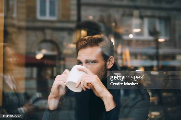 businessman drinking coffee while looking away in cafe seen through glass window - coffee drink photos et images de collection