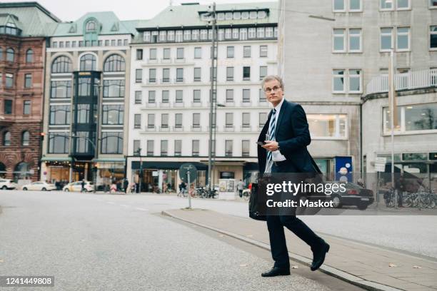 mature entrepreneur using smart phone while crossing road in city - überqueren stock-fotos und bilder