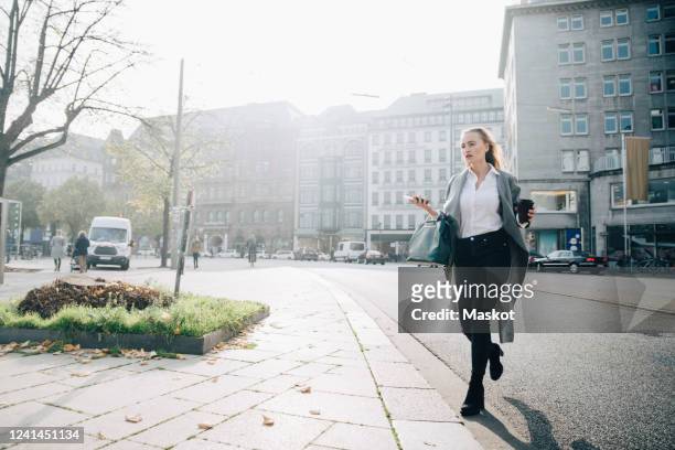 full length of female entrepreneur with smart phone walking in city - street worker stock-fotos und bilder