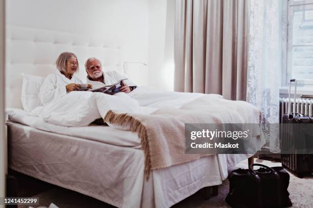 senior woman reading newspaper while having coffee by man relaxing on bed in hotel - paper city stock-fotos und bilder