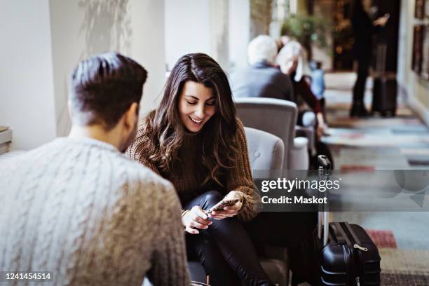 smiling woman using smart phone while sitting with male partner on chair in hotel - suitcase couple stock pictures, royalty-free photos & images