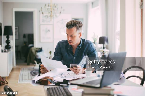 male architect sitting at workplace reading blueprint at home - mature reading computer bildbanksfoton och bilder