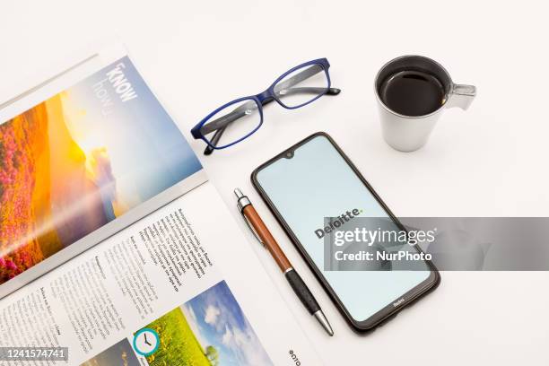 In this photo illustration a Deloitte logo seen displayed on a smartphone screen on a desk next to a cafe, a pen, glasses and a magazine in Athens,...