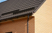A close-up on a metal tiled rooftop with roof snow guards, snow stoppers, and a rain gutter with a downspout on a brick house.