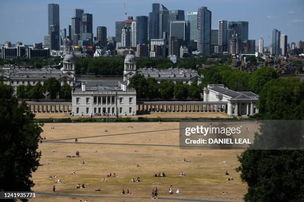 People sit on the sun-scorched grass near the Old Royal Naval College backdropped by the Canary Wharf financial district, in Greenwich Park, south...