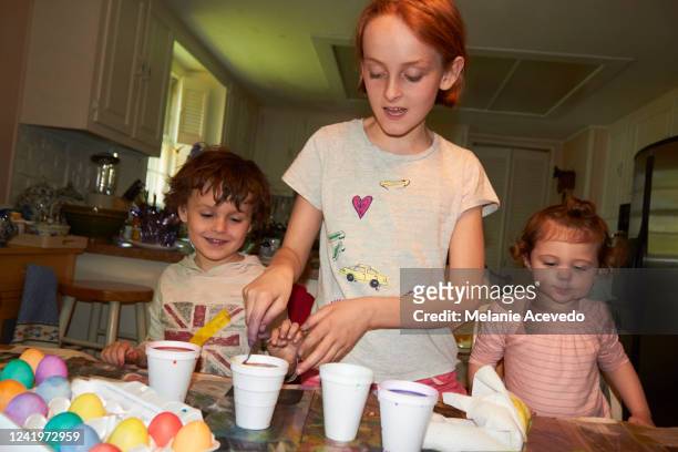 young cousins dying eggs for easter at kitchen table camera flash older redheaded girl standing up helping younger brother and cousin with easter tradition brother is laughing - children only stock-fotos und bilder
