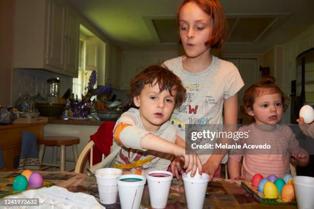 young cousins dying eggs for easter at kitchen table camera flash older redheaded girl standing up helping younger brother and cousin with easter tradition young boy with short brown curly hair is reaching over the table to the foam cup - melanie cousins stock-fotos und bilder