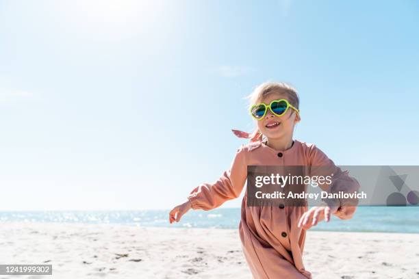 five years old little girl smiling and running on the beach in a sunny cloudless day. - blue sunglasses stock pictures, royalty-free photos & images