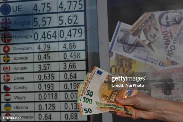 Person holds banknotes of the Euro in front of a board showing the selling and buying rates of various currencies in relation to the Polish Zloty. On...