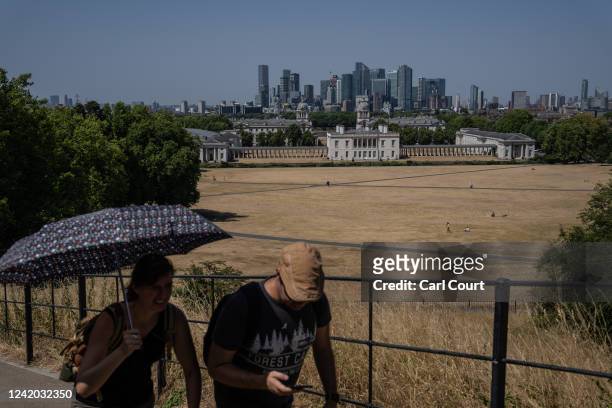 Woman shelters under an umbrella as she walks past dried grass in Greenwich Park on July 19, 2022 in London, England. Temperatures were expected to...