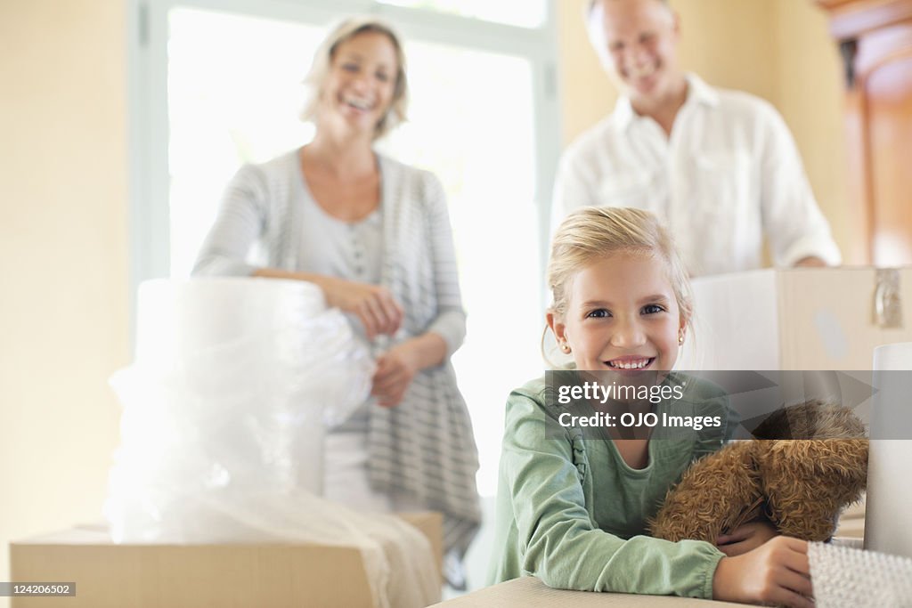 Portrait of smiling girl holding teddy bear with moving boxes, parents in background