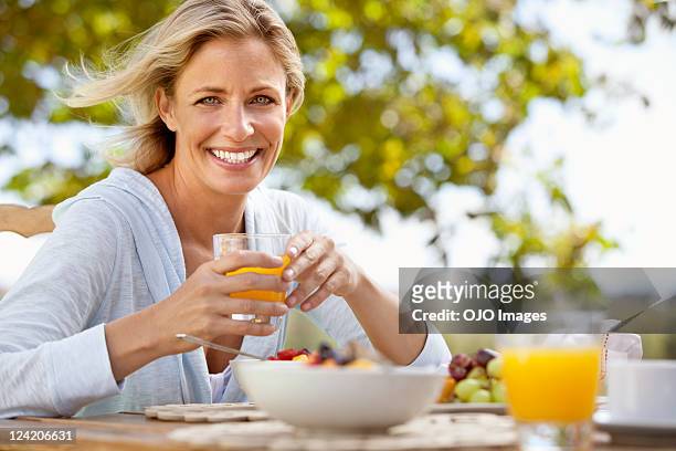 sorridente mulher madura com suco de laranja na mesa de pequeno-almoço outdoo - breakfast lifestyle female imagens e fotografias de stock