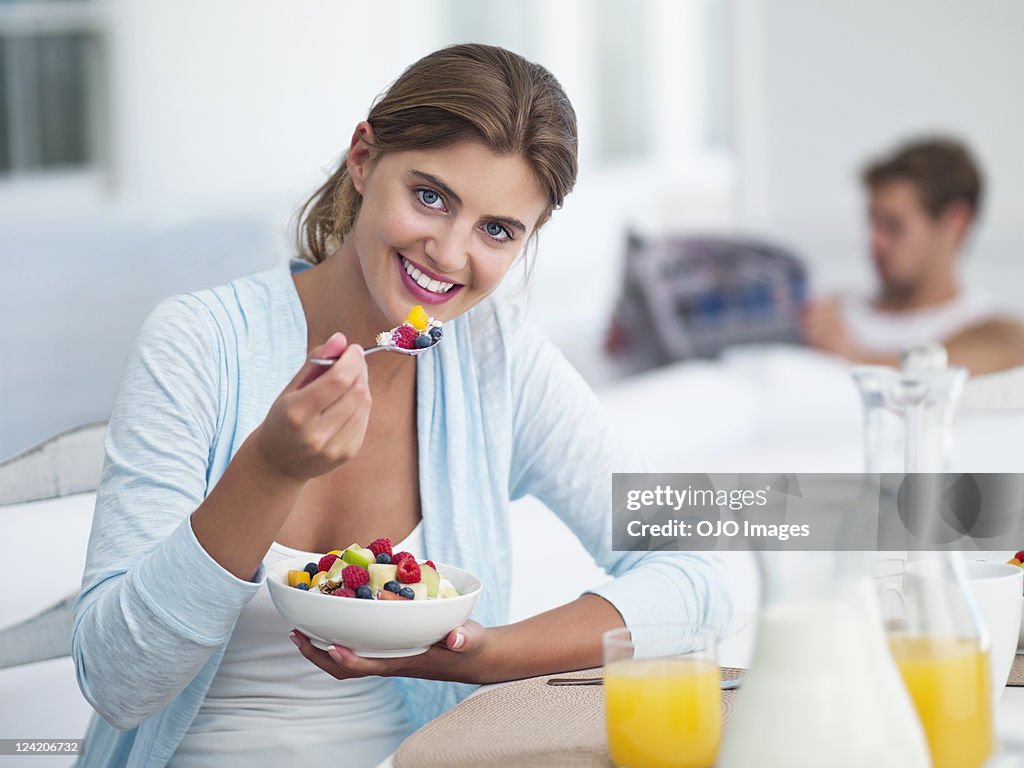 Young woman eating fruit salad with boyfriend in the background