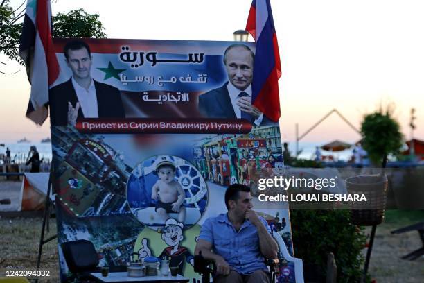 Man sits in front of a poster depicting Syrian President Bashar al-Assad and Russian President Vladimir Putin and reading in Arabic "Syria stands...