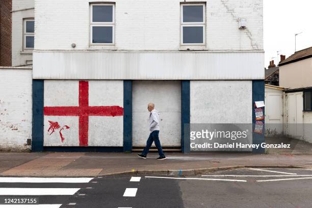 Man walks past a long-closed and boarded up shop whose front has been vandalised by a graffitied Cross of Saint George, the national flag of England,...