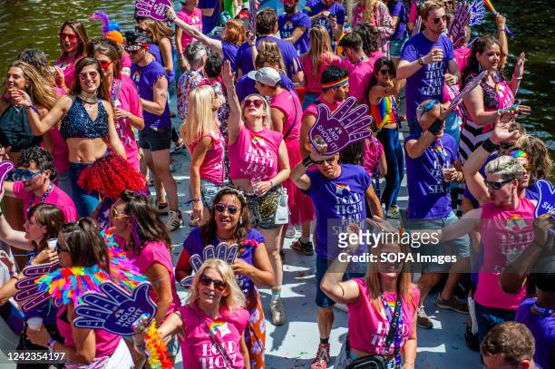 People wearing pink and purple clothes are seen cheering the audience from a boat. The Canal Parade is what Amsterdam Gay Pride is famous for. It's...