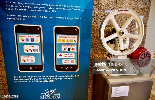 Tablet press, a mechanical device used to compress powder into tablets, is displayed next to a poster showing emojis used to sell drugs online, at...