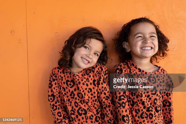 twin sisters with matching outfits laughing and standing in front of orange background - mesma roupa imagens e fotografias de stock