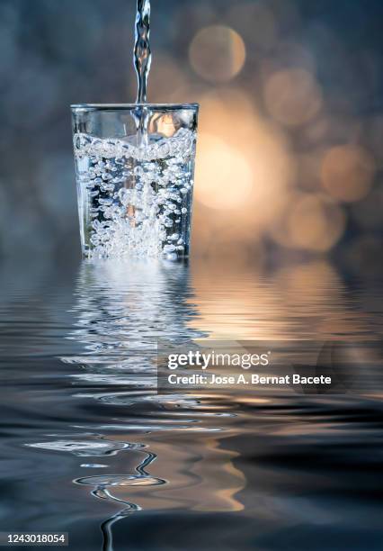 fill a glass of water that overflows, illuminated by sunlight and reflected on the water. - overflowing glass stockfoto's en -beelden