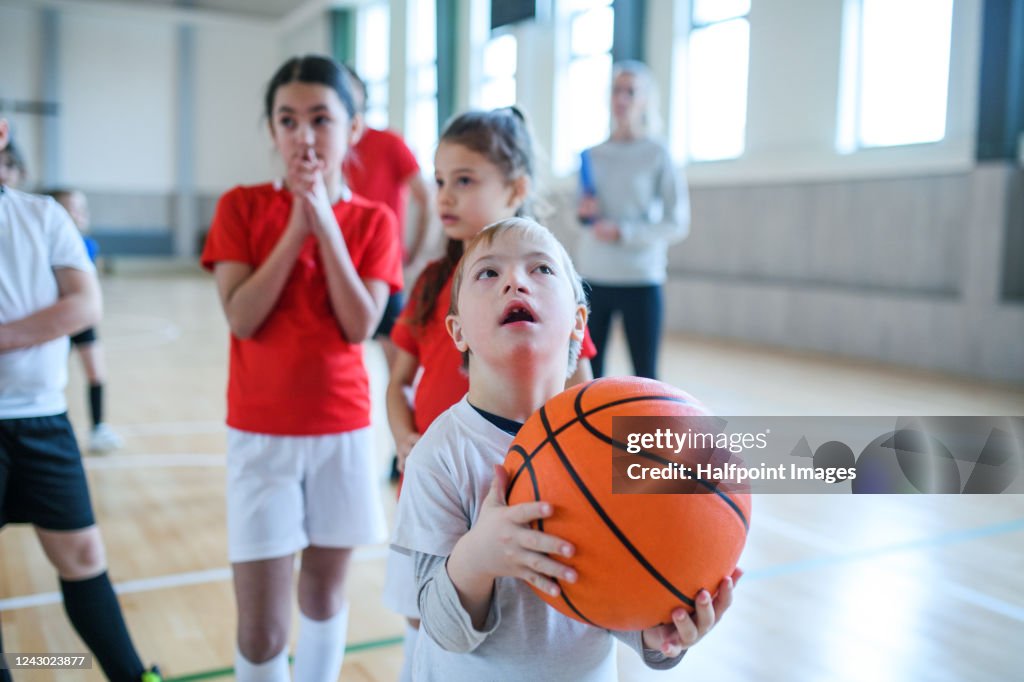 Down syndrome boy with pupils exercising indoors in gym class, playing basketball.