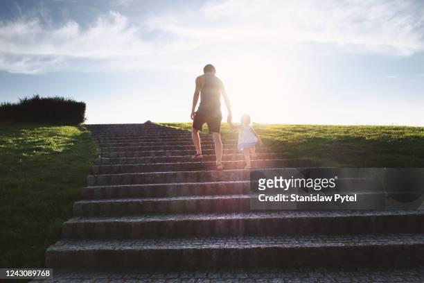 father wlaking with  daughter by hand on big stairs in park at susnet - family tree stockfoto's en -beelden