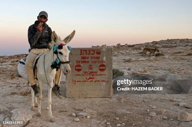 Palestinian man rides a donkey near a concrete sign showing a warning in Hebrew, Arabic, and English denoting a firing range zone in the southern...