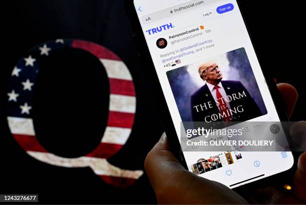 In this photo illustration, Donald Trump's TRUTH Social account is seen on a mobile device with an image of a QAnon sticker in the background in...