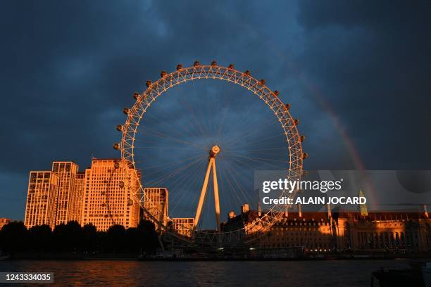 The evening sunlight falls on the London Eye as a rainbow is seen in the sky, nearby, members of the public queue on the South Bank to pay their...