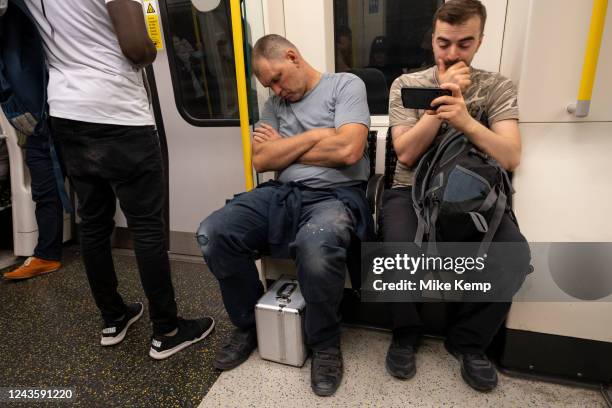 Two men with different ways to spend their time on a London Underground train, with one sleeping while the other looks at content on his smartphone...