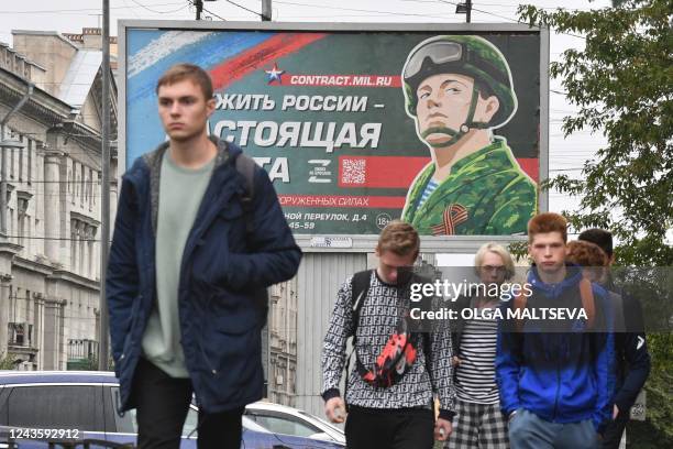 Young men walk in front of a billboard promoting contract army service with an image of a serviceman and the slogan reading "Serving Russia is a real...