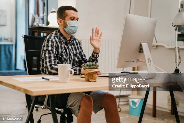young adult man is having a work video call with a desktop pc - makeshift desk stock pictures, royalty-free photos & images