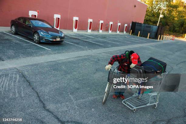 Cedric Thompkins, homeless since June 2022, pulls his possessions in a shopping cart past a car at a Tesla charging station in Bloomington. Thompkins...