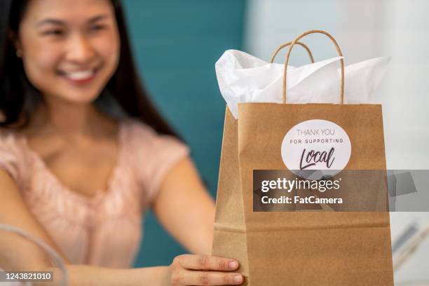 sonriente mujer propietaria de la tienda sosteniendo una bolsa de artesanía con una etiqueta adhesiva "gracias por apoyar a local" - tienda de regalos fotografías e imágenes de stock